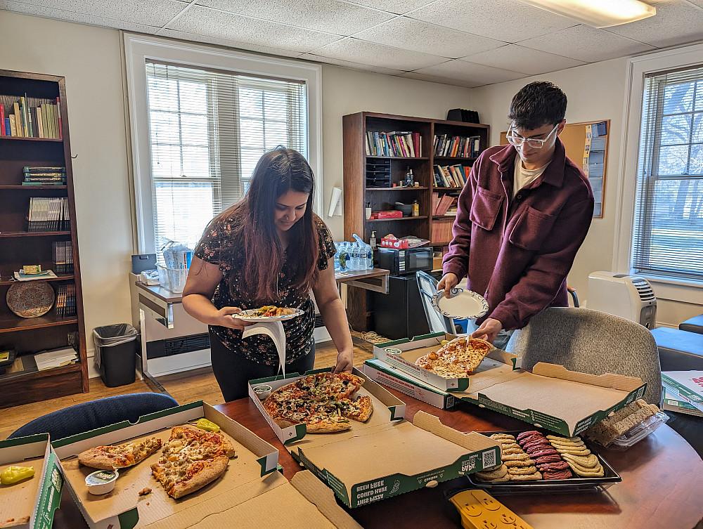 two students stand by a table reaching for pizza