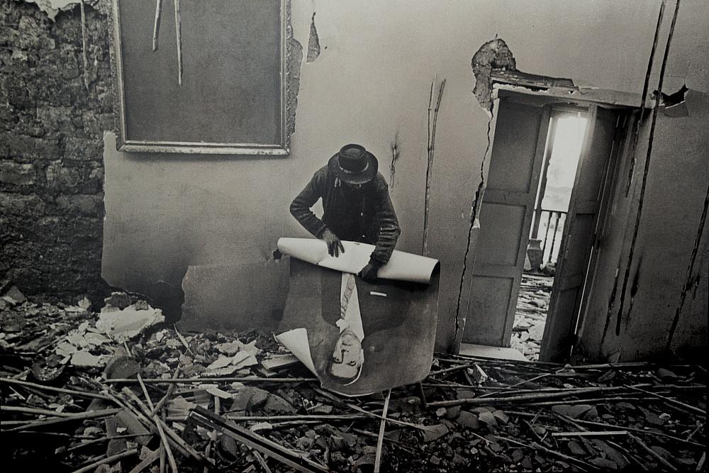 A victim of violence rolls up a poster in the midst of rubble.